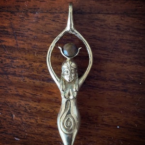 K&ouml;nnte beinhalten: Ein silberner Anh&auml;nger mit einer stilisierten weiblichen Figur mit erhobenen Armen, die einen braunen Edelstein h&auml;lt. Die Figur hat ein Spiralmuster am unteren K&ouml;rper.