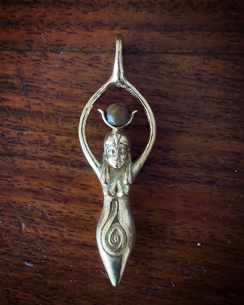 K&ouml;nnte beinhalten: Ein silberner Anh&auml;nger mit einer stilisierten weiblichen Figur mit erhobenen Armen, die einen braunen Edelstein h&auml;lt. Die Figur hat ein Spiralmuster am unteren K&ouml;rper.