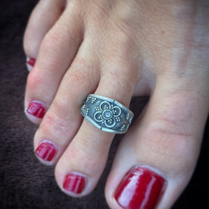 May include: A silver toe ring with a floral design. The ring is worn on a toe with red nail polish.