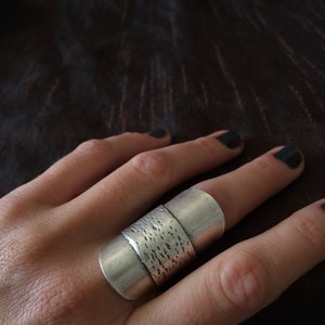 May include: A silver ring with a textured band and two smooth bands. The ring is worn on a finger. The ring has a modern, minimalist design.