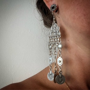 Può includere: Orecchini pendenti in argento con una parte superiore decorativa e più catene con ciondoli a forma di moneta.