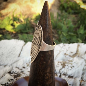 May include: A silver-colored ring with a detailed wing design. The ring is displayed on a dark wooden cone-shaped stand against a blurred background of greenery and sunlight. The wing has intricate feather detailing.