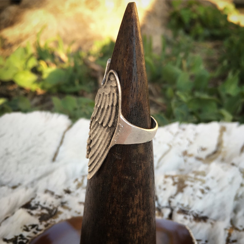 May include: A silver-colored ring with a detailed wing design. The ring is displayed on a dark wooden cone-shaped stand against a blurred background of greenery and sunlight. The wing has intricate feather detailing.