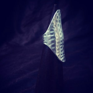 May include: A silver ring with a textured, hammered design. The ring is displayed on a black stand.