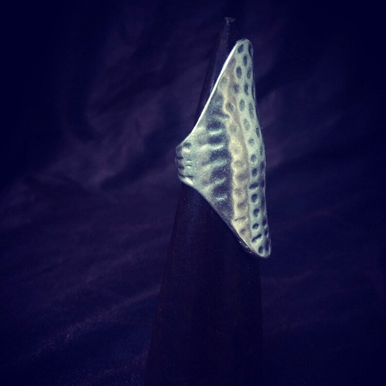 May include: A silver ring with a textured, hammered design. The ring is displayed on a black stand.