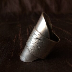 May include: A silver-colored ring with a textured band and a pointed, elongated design. The ring has a slightly tarnished appearance, suggesting an antique or vintage style. The ring is set against a dark brown background.