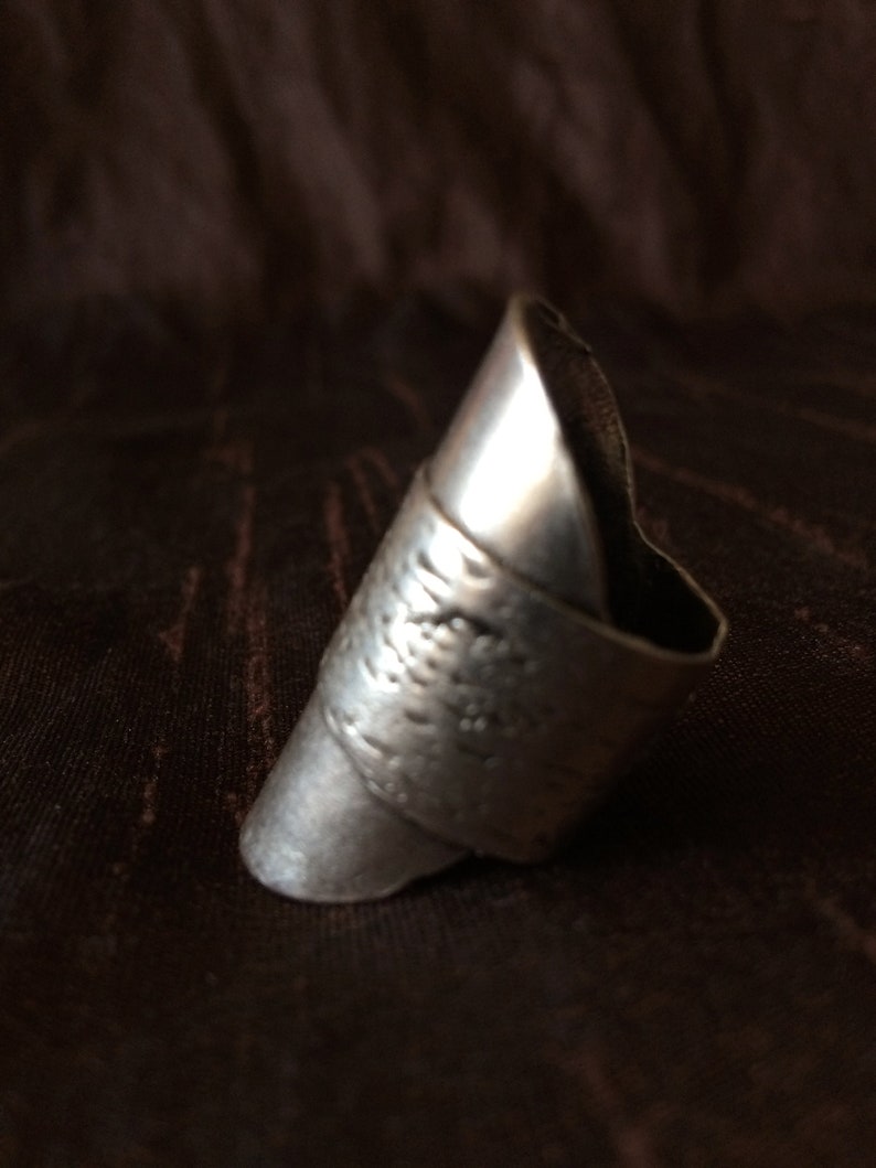 May include: A silver-colored ring with a textured band and a pointed, elongated design. The ring has a slightly tarnished appearance, suggesting an antique or vintage style. The ring is set against a dark brown background.