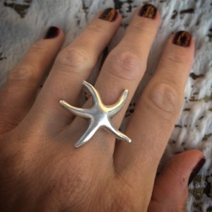 Può includere: Un anello a forma di stella marina in argento con una superficie testurizzata. L'anello è indossato sul dito medio di una mano.