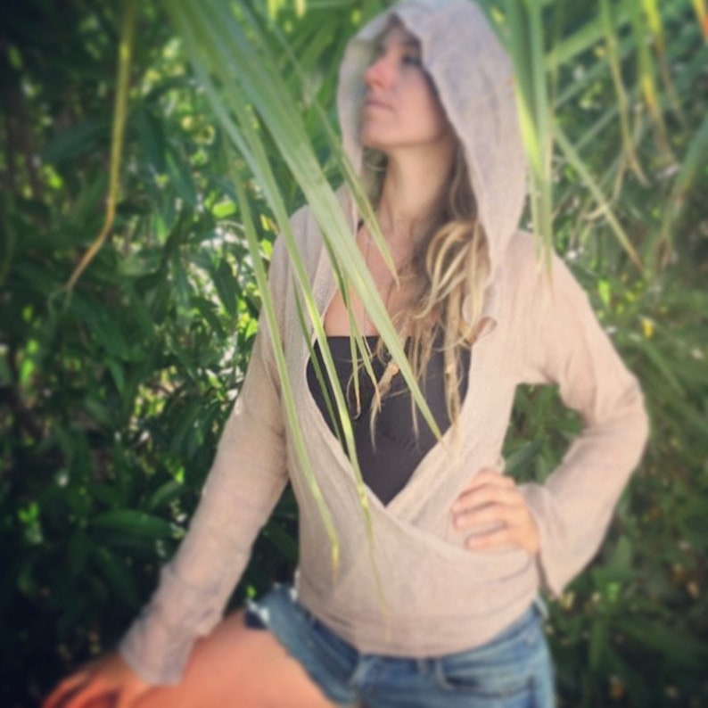 May include: A woman wearing a light beige hooded cardigan with a black tank top underneath. The cardigan is open in the front, revealing the tank top. The woman has long, blonde hair and is standing in front of a green leafy background.