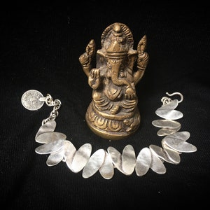 Può includere: Un bracciale in argento con maglie a forma di foglia e un piccolo ciondolo a forma di moneta. Una statua in bronzo della divinità indù Ganesha è posizionata dietro il bracciale. Su sfondo nero.