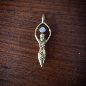 K&ouml;nnte beinhalten: Ein goldener Anh&auml;nger mit einer stilisierten Frauenfigur, die eine wei&szlig;e Steinkugel &uuml;ber ihrem Kopf h&auml;lt. Die Figur hat ein Spiralmuster auf ihrem Oberk&ouml;rper.