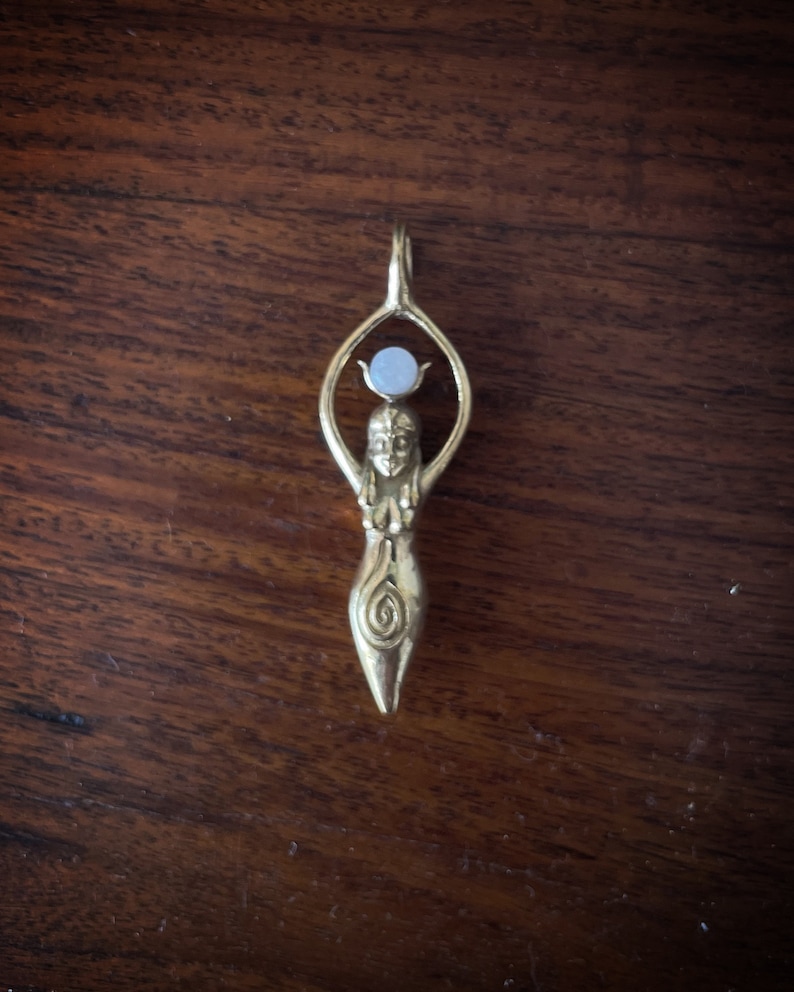 K&ouml;nnte beinhalten: Ein goldener Anh&auml;nger mit einer stilisierten Frauenfigur, die eine wei&szlig;e Steinkugel &uuml;ber ihrem Kopf h&auml;lt. Die Figur hat ein Spiralmuster auf ihrem Oberk&ouml;rper.