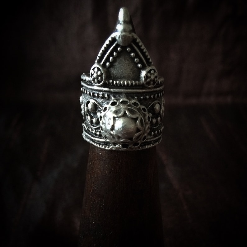 May include: A silver ring with a detailed, ornate design. The ring features a pointed top and a circular center piece with intricate patterns.