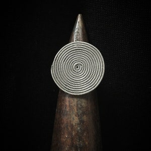 May include: A silver spiral ring with a circular design. The ring is displayed on a wooden stand.