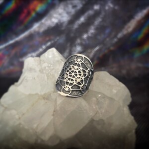 Può includere: Un anello argentato con un intricato design geometrico. L'anello ha una forma rettangolare con bordi arrotondati e un motivo dettagliato di linee e forme interconnesse. È esposto su una superficie cristallina bianca.