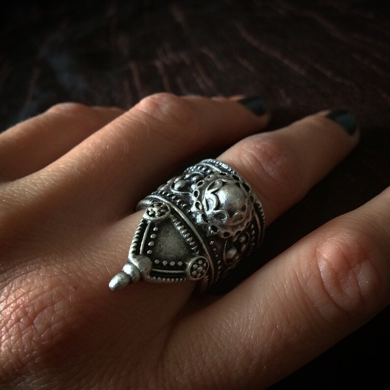 May include: A silver ring with intricate details and a textured design. The ring is wide and has a pointed top.