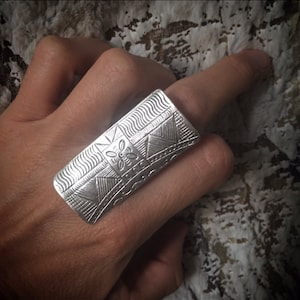 Può includere: Un anello d'argento con un design geometrico. L'anello è largo e piatto, e ha una superficie testurizzata.