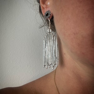 Puede incluir: Pendientes de plata con diseño geométrico. Los pendientes están hechos de finas líneas verticales que crean una forma rectangular. Los pendientes cuelgan de un pendiente de plata.