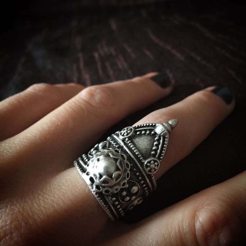 May include: A silver ring with intricate designs and a raised, rounded top. The ring is worn on a finger.