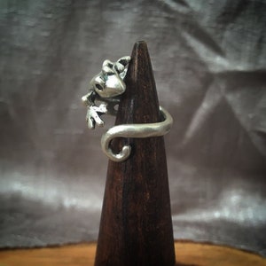 May include: A silver ring featuring a detailed gecko design. The gecko is curled around the band of the ring, with its tail wrapping around the finger.
