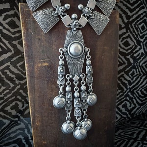 Può includere: Una collana d'argento con un grande ciondolo decorato e più ciondoli pendenti. Il ciondolo ha la forma di un diamante e presenta dettagli intricati. I ciondoli sono rotondi e hanno una superficie leggermente testurizzata.