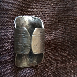May include: A silver-toned ring with a textured band and a wide, curved face. The ring has a rustic, hammered appearance, suggesting a handcrafted design. The metal has a slightly tarnished look, adding to its vintage appeal.