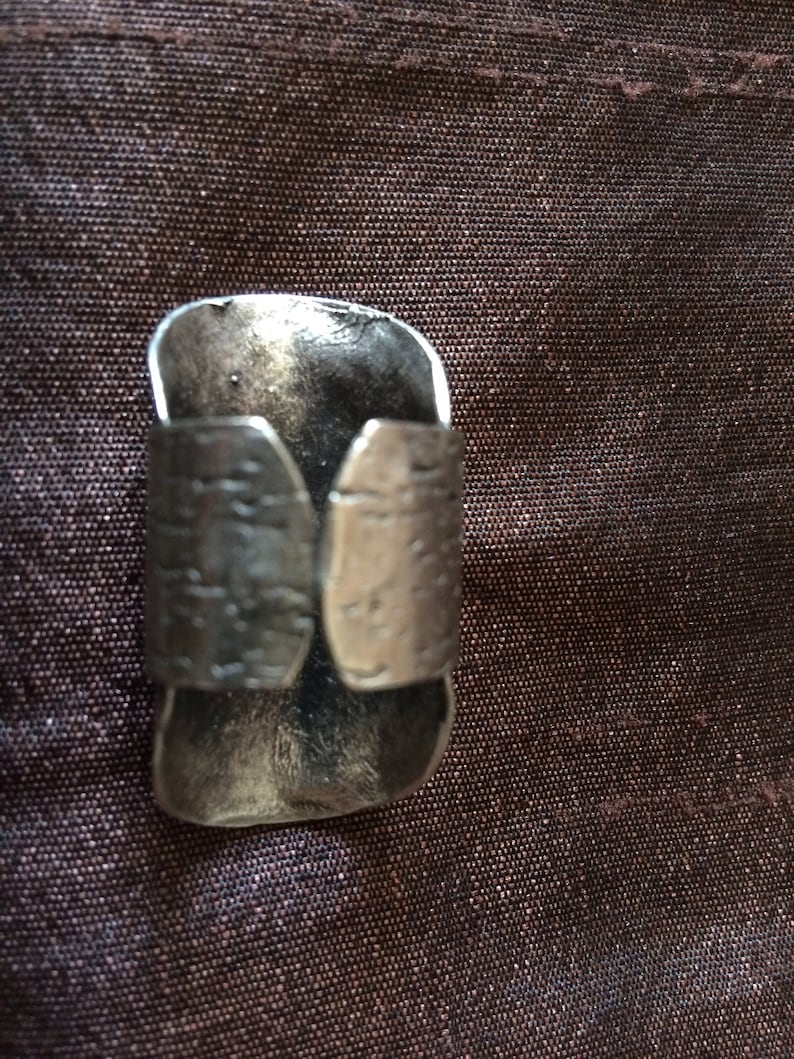 May include: A silver-toned ring with a textured band and a wide, curved face. The ring has a rustic, hammered appearance, suggesting a handcrafted design. The metal has a slightly tarnished look, adding to its vintage appeal.