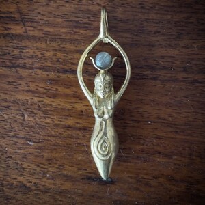 K&ouml;nnte beinhalten: Goldener Anh&auml;nger mit einer stilisierten weiblichen Figur mit erhobenen Armen und einem runden Edelstein an der Spitze. Die Figur hat ein detailliertes Design mit einem Spiralmuster am Boden.