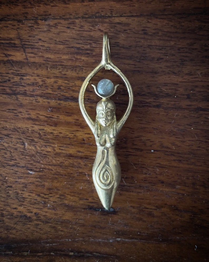 K&ouml;nnte beinhalten: Goldener Anh&auml;nger mit einer stilisierten weiblichen Figur mit erhobenen Armen und einem runden Edelstein an der Spitze. Die Figur hat ein detailliertes Design mit einem Spiralmuster am Boden.