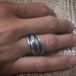 Può includere: Un anello d'argento con un designo intrecciato. L'anello è indossato su un dito.