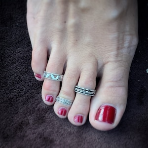 Può includere: Tre anelli per dita dei piedi in argento con diversi design. Gli anelli sono indossati sul dito grosso del piede, sul secondo dito del piede e sul terzo dito del piede. Le dita dei piedi sono dipinte con smalto rosso.