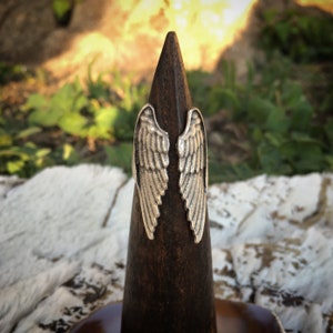 May include: A pair of silver angel wings attached to a dark brown wooden ring holder.