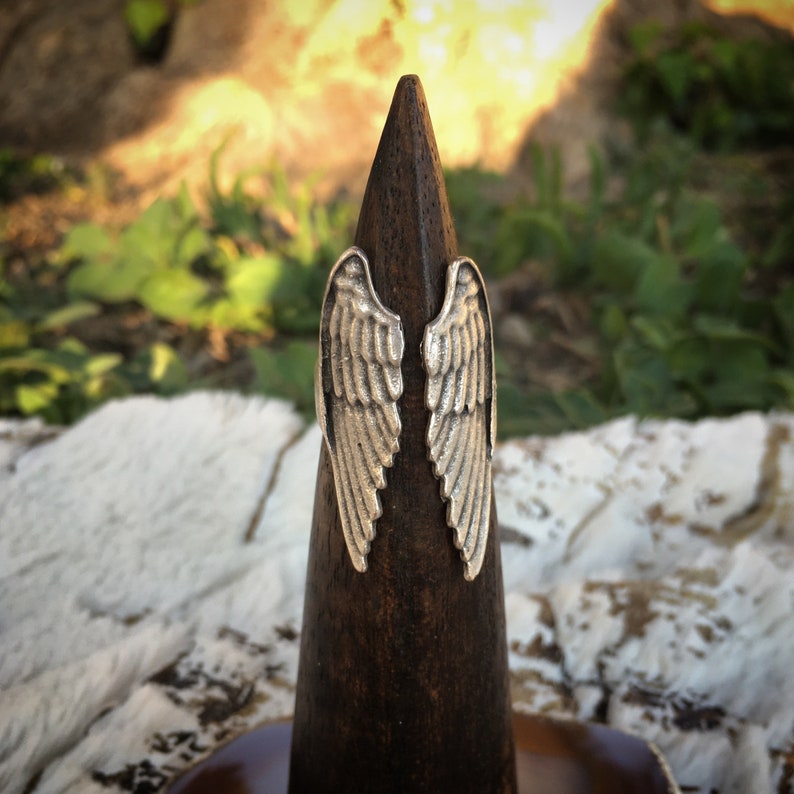 May include: A silver-colored ring with an angel wing design. The ring is displayed on a dark brown wooden cone-shaped stand. The wings are detailed with individual feather lines.