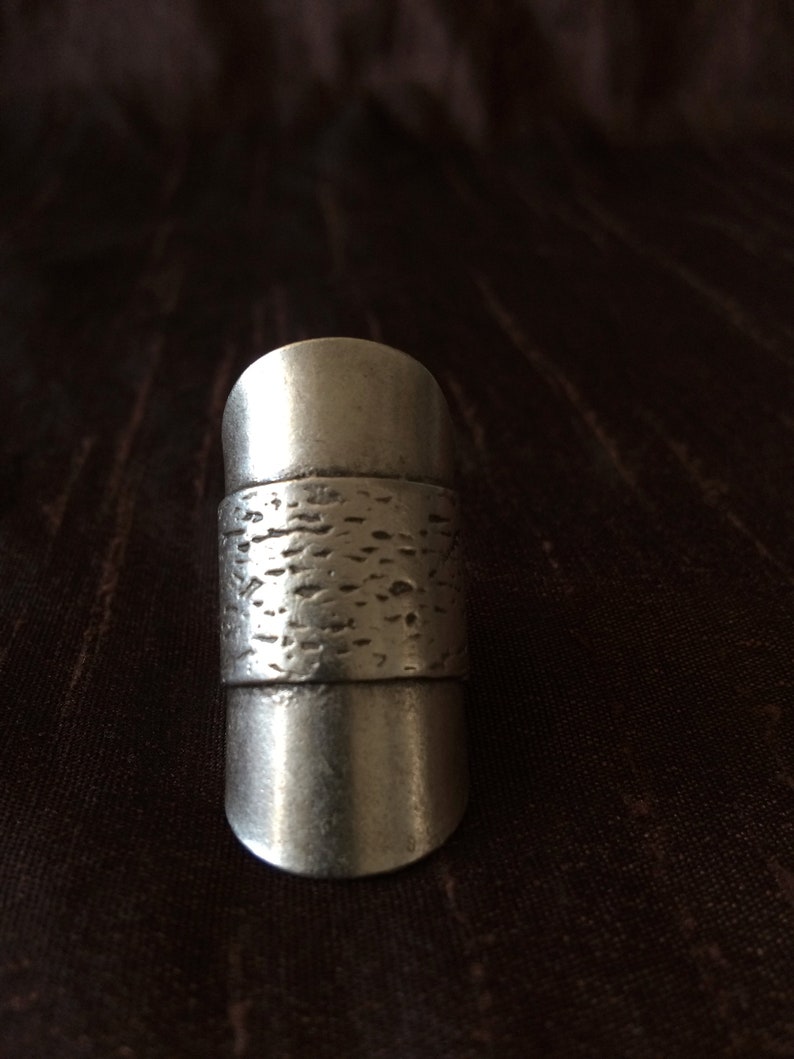May include: A silver-colored ring with a textured band. The ring has a cylindrical shape with a smooth, rounded top and bottom. The textured band adds a unique detail to the otherwise simple design.