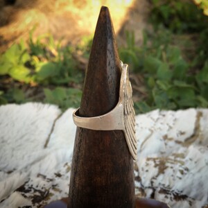 May include: Silver-toned ring with a detailed wing design. The ring is displayed on a dark brown wooden cone against a backdrop of green foliage and a white, textured surface. The wing design is the focal point, showcasing intricate feather detailing.