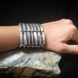Puede incluir: Un brazalete de metal plateado con un diseño corrugado. El brazalete es ancho y tiene una superficie texturizada.