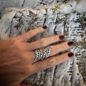 May include: A silver ring with a leaf design. The ring is worn on the middle finger of a hand with dark red nail polish.