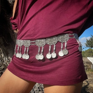 Puede incluir: Un cinturón de monedas de plata con una parte superior burdeos. El cinturón está hecho de una cadena con colgantes decorativos en forma de monedas.