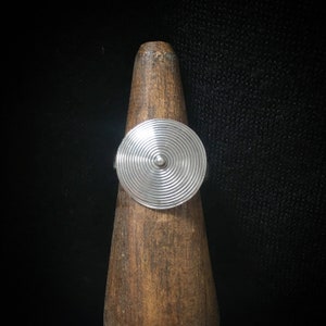 May include: A silver ring with a circular design featuring concentric circles. The ring is displayed on a wooden stand.