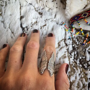 May include: A silver-toned ring with a wing design. The ring features two detailed wing shapes, each with etched feather patterns. The ring is worn on a finger, set against a textured white and brown background.