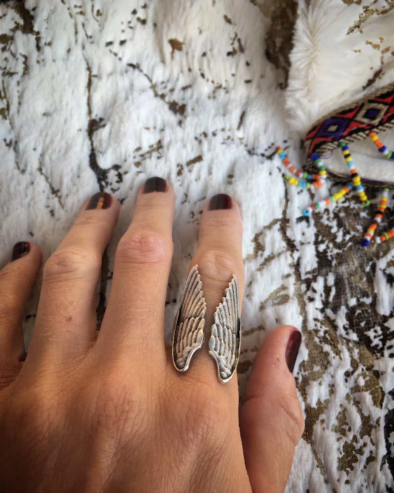 May include: A silver-toned ring with a wing design. The ring features two detailed wing shapes, each with etched feather patterns. The ring is worn on a finger, set against a textured white and brown background.