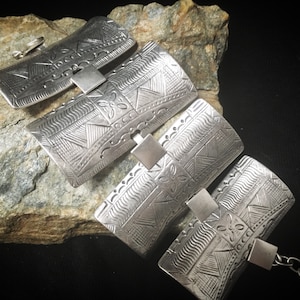 Può includere: Un braccialetto d'argento con un motivo geometrico. Il braccialetto è composto da più sezioni collegate, ciascuna con un design diverso. Il braccialetto è su una roccia grigia.