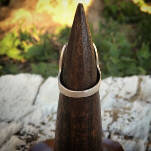 May include: A silver-colored ring with a curved design is displayed on a dark brown wooden cone. The ring has a smooth, polished surface and a simple, elegant style. The background is blurred, with hints of green foliage and sunlight.