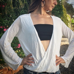 Puede incluir: Una blusa blanca de manga larga, estilo cruzado, con un bandeau negro debajo. La blusa tiene un escote en V profundo y está hecha de una tela ligera y fluida.