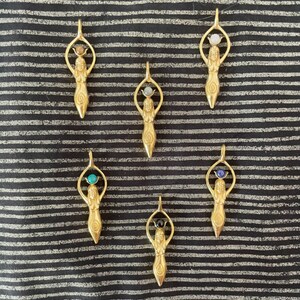 K&ouml;nnte beinhalten: Sechs goldene Anh&auml;nger mit einer weiblichen Figur, die einen Edelstein h&auml;lt. Die Edelsteine sind in verschiedenen Farben, darunter blau, gr&uuml;n, schwarz und wei&szlig;.