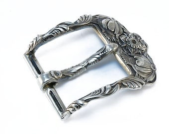 May include: A silver belt buckle with an ornate floral design. The buckle is made of metal and has a polished finish.