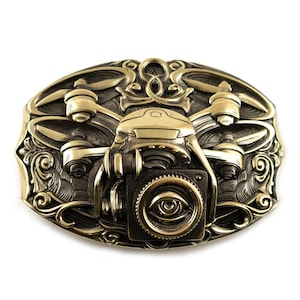 May include: A gold-toned belt buckle with an intricate design featuring a stylized drone with a camera lens. The camera lens has an eye-like design.