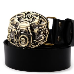 Puede incluir: Un cinturón de cuero negro con una hebilla de color latón antiguo. La hebilla presenta un diseño steampunk con engranajes, un ojo y elementos mecánicos. El cinturón mide aproximadamente 3,8 cm de ancho.