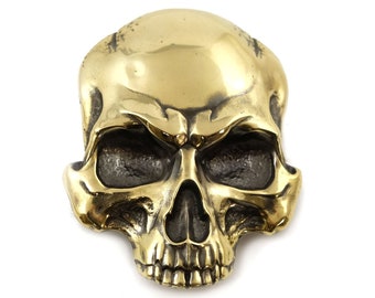 Puede incluir: Una hebilla de cinturón de calavera de color dorado con un diseño detallado. La calavera tiene una frente prominente y una mandíbula definida.