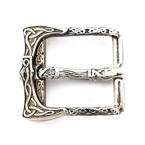 May include: Silver belt buckle with a Celtic knot design and an axe head shape. The buckle is detailed with intricate carvings and has a polished finish.